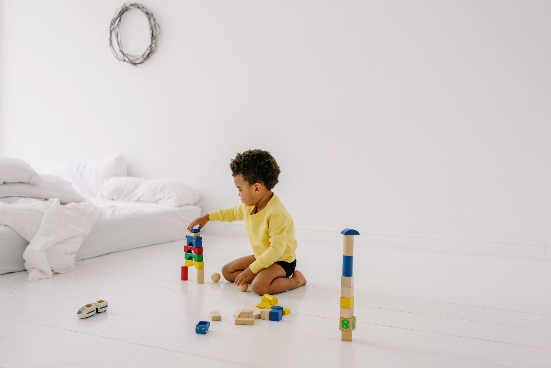 Young child in yellow plays with colorful wooden blocks on a white bedroom floor.