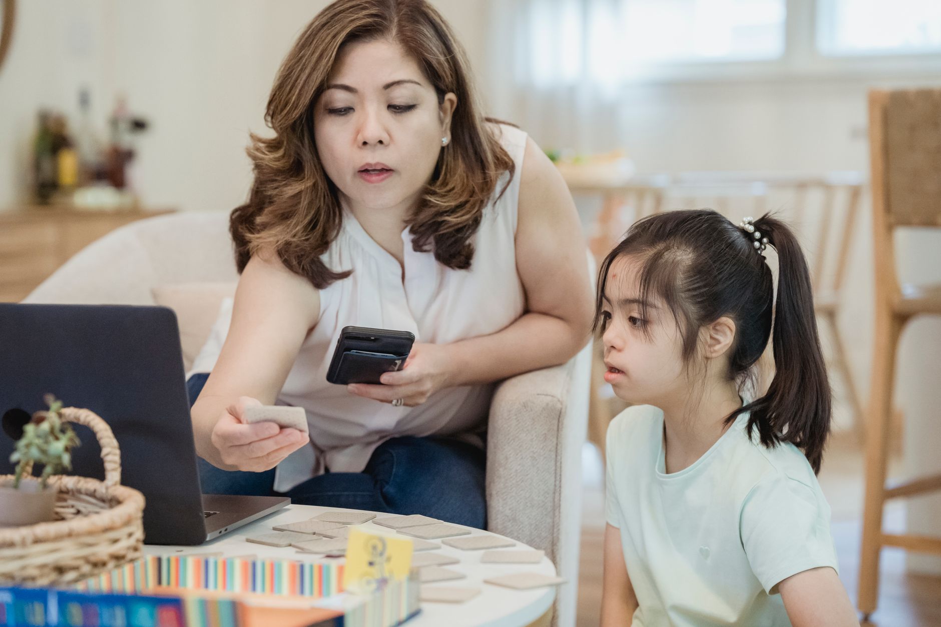 A mother guides her daughter in learning activities using cards and a laptop at home.